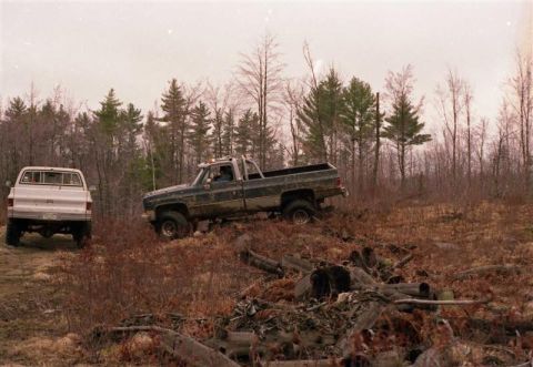 1991 Ford Bronco - Two pickup trucks are seen off-roading in a wooded area with fallen logs and brush.