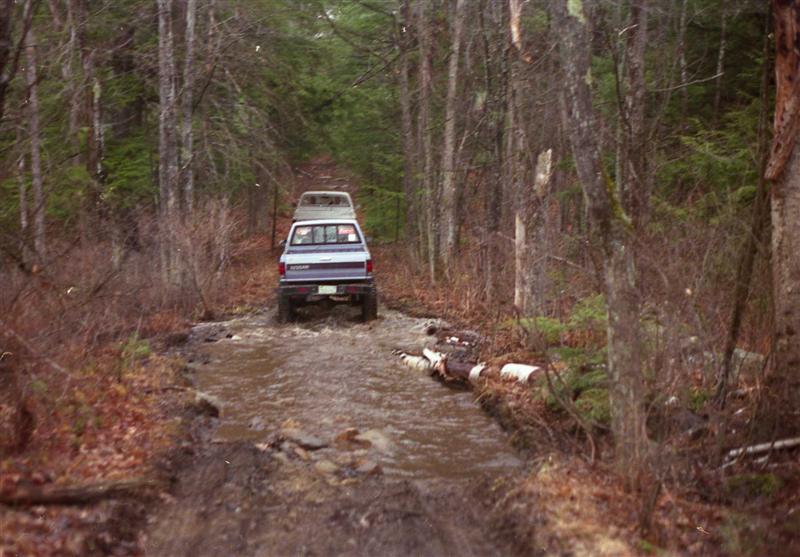 Rear view of a pickup truck driving through a muddy forest path, another truck follows
