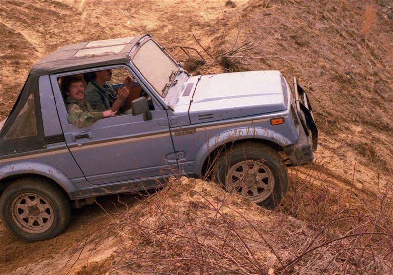 Suzuki Samurai with two people inside navigating a dirt hill, front wheel deep in mud