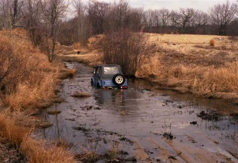 1991 Ford Bronco - A Suzuki Samurai navigates a water-filled trail surrounded by dry brush and trees.