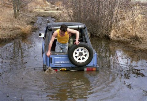 1991 Ford Bronco - A man attempts to exit a blue Suzuki Samurai that is stuck in a muddy, watery trail.
