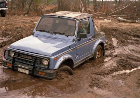 1991 Ford Bronco - A blue Suzuki Samurai is deeply stuck in thick mud, with another vehicle visible in the background.