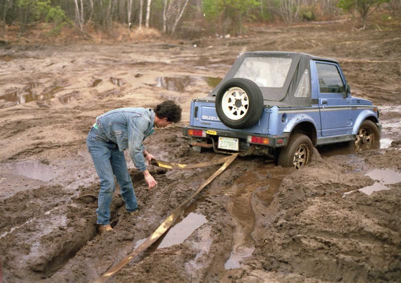 Blue Suzuki Samurai stuck in mud, person trying to free it with wood planks. License plate 227981