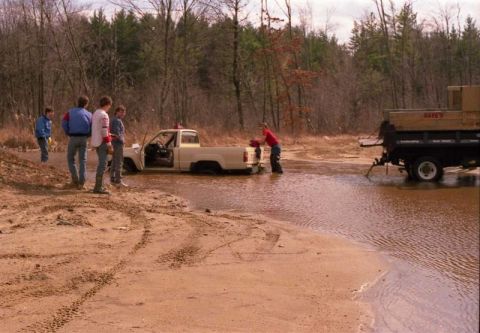 1991 Ford Bronco - A group of people watch as a pickup truck is stuck in a muddy water crossing, with a trailer nearby.