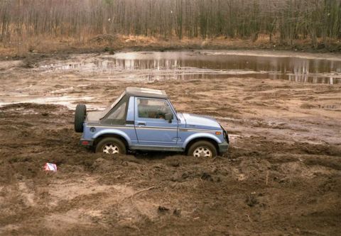 1991 Ford Bronco - A blue Suzuki Samurai is stuck deep in the mud, with only its tires and lower body visible.