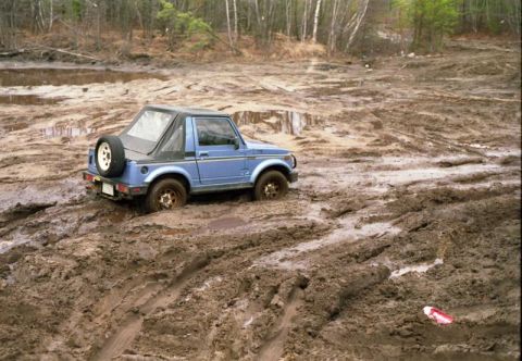 1991 Ford Bronco - Suzuki Samurai struggling in muddy terrain with trees in the background.