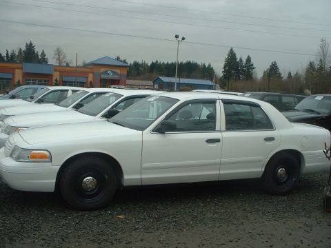 1999 Ford Crown Victoria - A row of white sedans, likely police cruisers, are parked outside a commercial building under an overcast sky.