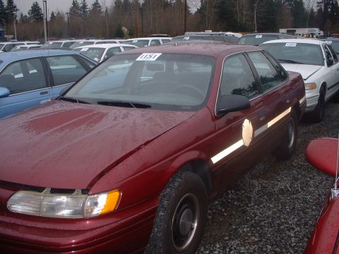1999 Ford Crown Victoria - A maroon sedan is parked in an outdoor lot with other vehicles, showing a reflective stripe and emblem on its side.