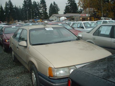 1999 Ford Crown Victoria - A tan sedan with a blue trim is parked in a lot with other vehicles under an overcast sky.