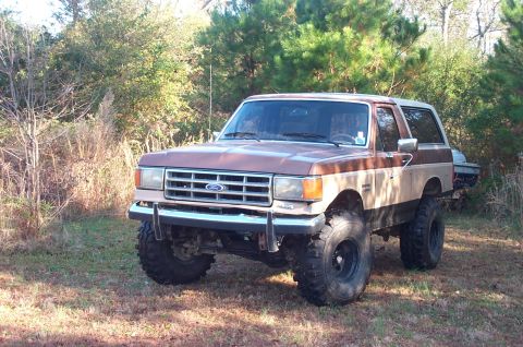 1987 Ford Bronco - A lifted 1987 Ford Bronco with two-tone paint sits in a grassy, wooded area.