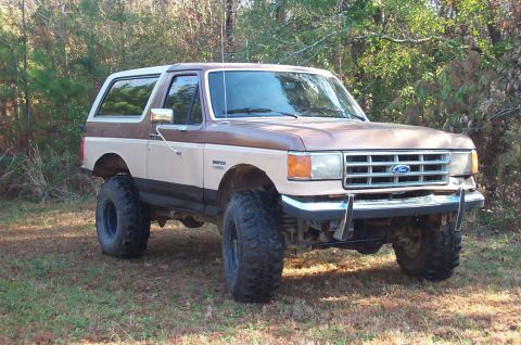 1987 Ford Bronco - This lifted 1987 Ford Bronco features a two-tone brown and tan paint scheme and is parked in a grassy, wooded area.
