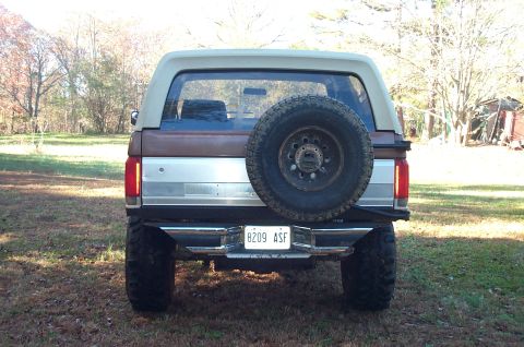 1987 Ford Bronco - Rear view of a brown and tan Ford Bronco with a spare tire on the back, parked outdoors.