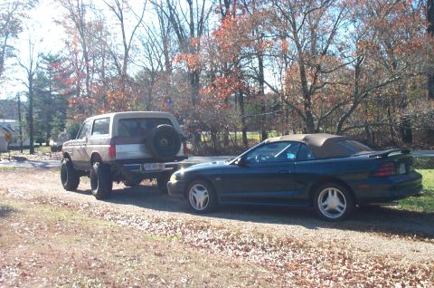 1987 Ford Bronco - A lifted Ford Bronco is parked on a dirt road, towing a convertible sports car.