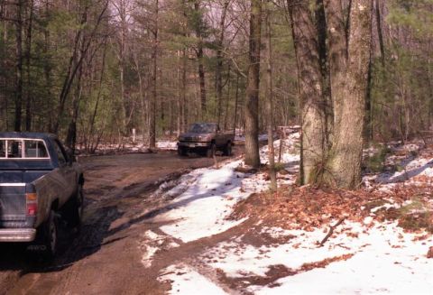 1991 Ford Bronco - Two trucks navigate a muddy and snowy path through a wooded area.