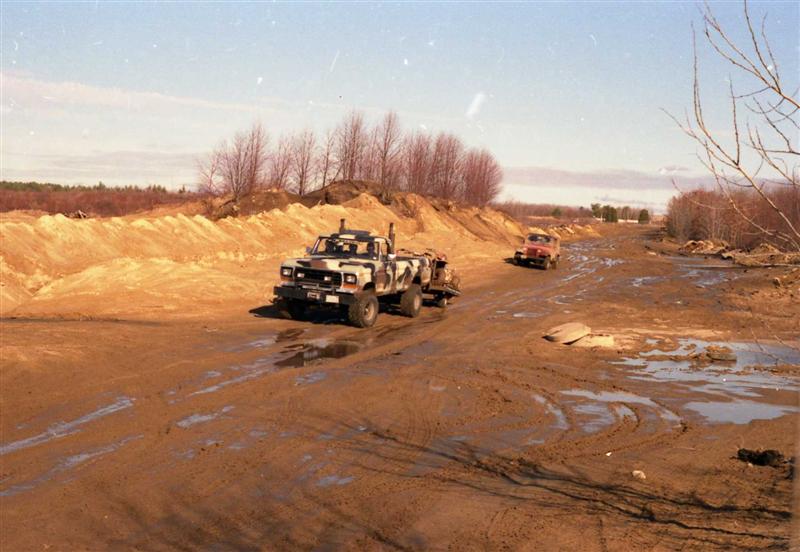 Camouflaged pickup truck with dual rear wheels towing a trailer on a muddy dirt road
