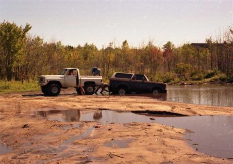 1991 Ford Bronco - A white pickup truck tows a dark SUV out of a muddy water hole, with trees in the background.
