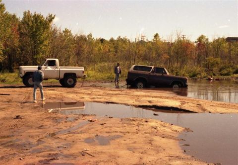 1991 Ford Bronco - Two vehicles are stuck in the mud and water, with trees and brush in the background.