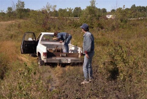 1991 Ford Bronco - Two individuals work with a winch in the bed of a white pickup truck that is stuck in a muddy, overgrown area.