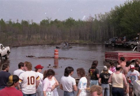 1991 Ford Bronco - A muddy scene with several off-road vehicles and spectators gathered around a water-filled track.