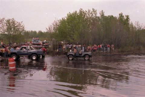 1991 Ford Bronco - Off-road vehicles traverse a muddy track as a crowd watches from the bank.
