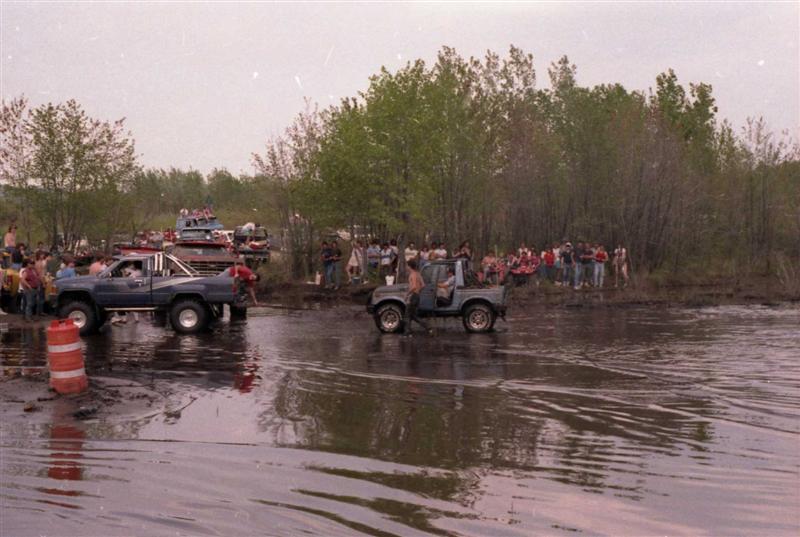 Side view of a blue pickup truck and a small gray SUV navigating a muddy area with onlookers