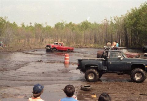 1991 Ford Bronco - Several trucks are navigating a muddy track with spectators watching from the side.