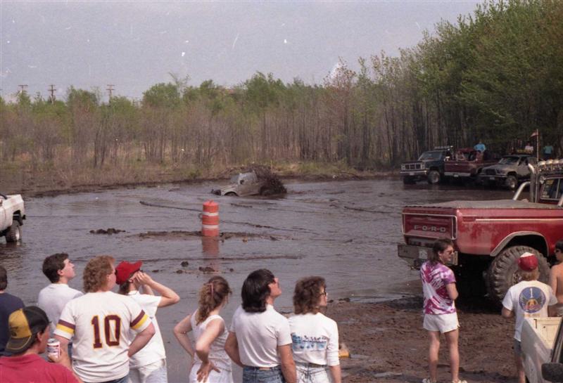 People watching trucks in a muddy field, one truck is stuck in the mud