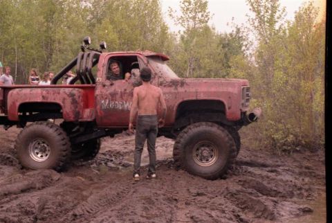 1991 Ford Bronco - A heavily modified red pickup truck is stuck in the mud with people around.