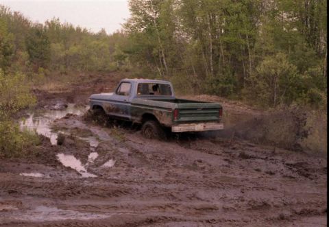 1991 Ford Bronco - A blue pickup truck with a green bed is stuck in deep mud on a wooded trail.