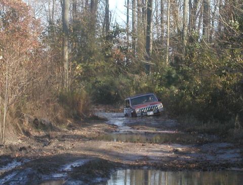 1988 Ford Bronco - A red Jeep Cherokee is stuck in deep mud on a wooded trail.