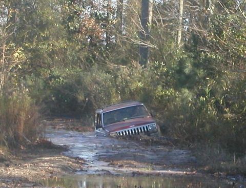 1988 Ford Bronco - A red Jeep Cherokee is stuck in a muddy trail, surrounded by dense vegetation.