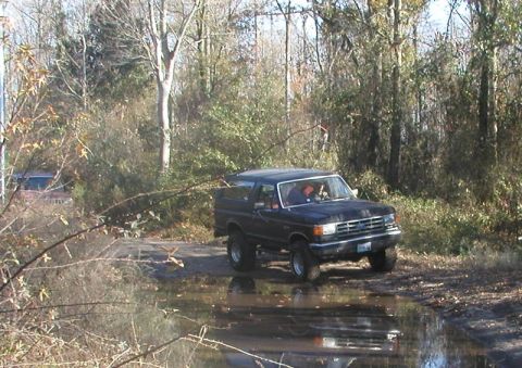1988 Ford Bronco - A black SUV with a lifted suspension navigates through a muddy, water-filled path in a wooded area.