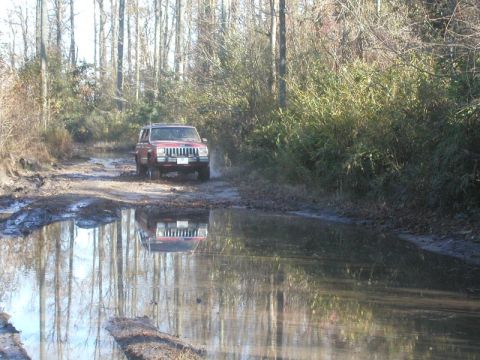 1988 Ford Bronco - Jeep Cherokee XJ navigates a muddy trail, its reflection visible in the water.