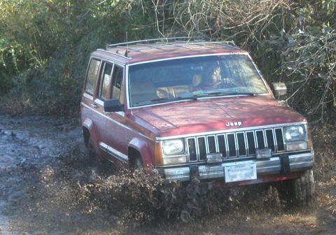 1988 Ford Bronco - A red Jeep Cherokee XJ navigates a muddy trail, kicking up a significant amount of dirt and water.