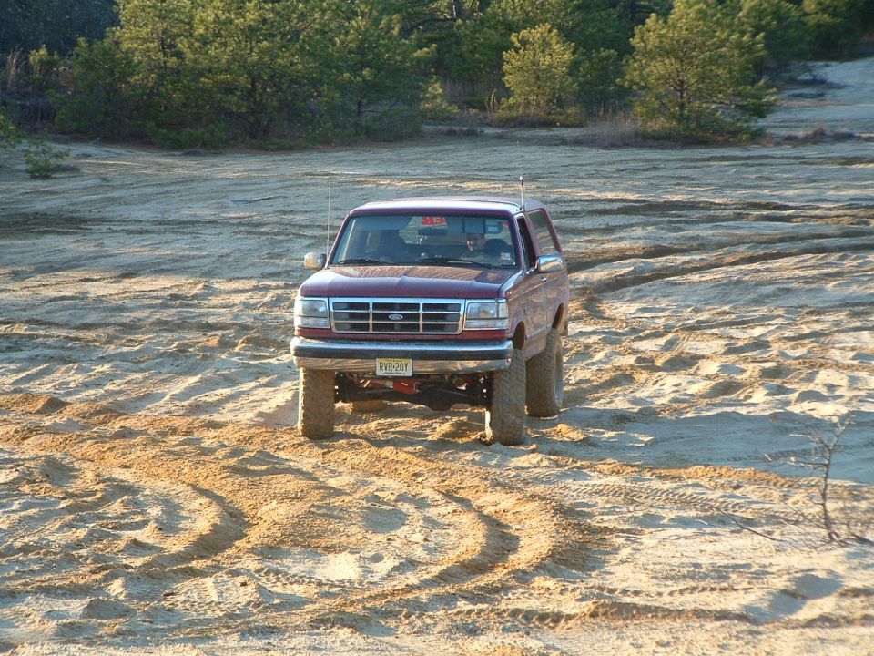 Maroon Ford Bronco driving on sandy terrain with trees in background. License plate reads RYY-20Y