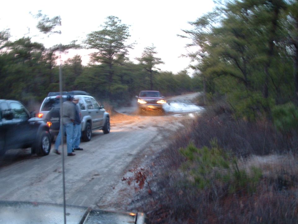Truck with headlights on splashing through a water hole on a sandy pine-woods trail as onlookers watch