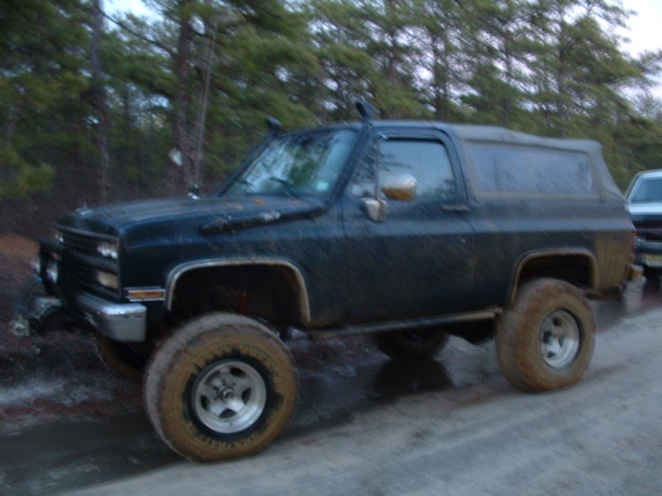Lifted dark blue two-door Chevy Blazer with muddy off-road tires on a wet dirt road