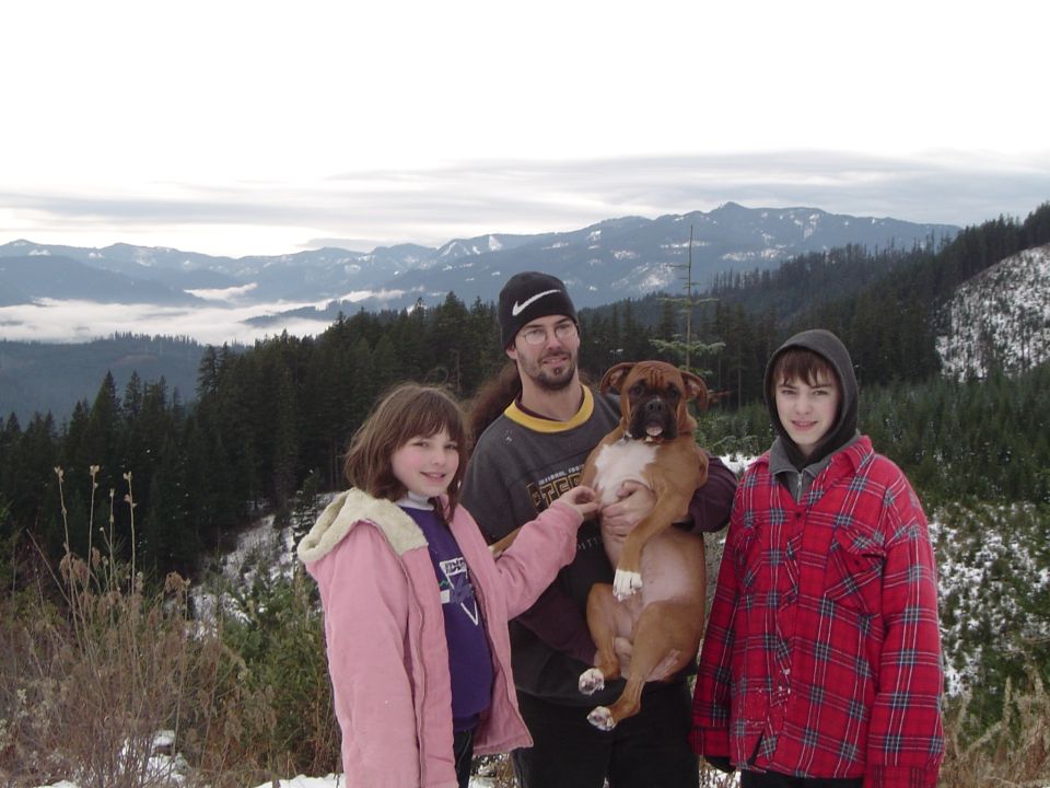 Man and two kids posing with a Boxer dog on a snowy mountain overlook with peaks behind