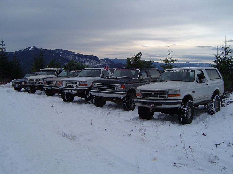 Row of lifted Ford trucks and Broncos parked in snow at dusk with mountains and a US flag