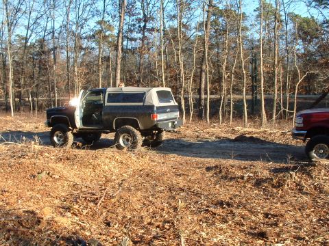1994 Ford Bronco - A black SUV with a tan soft top sits in a clearing surrounded by trees, with another vehicle partially visible to the right.