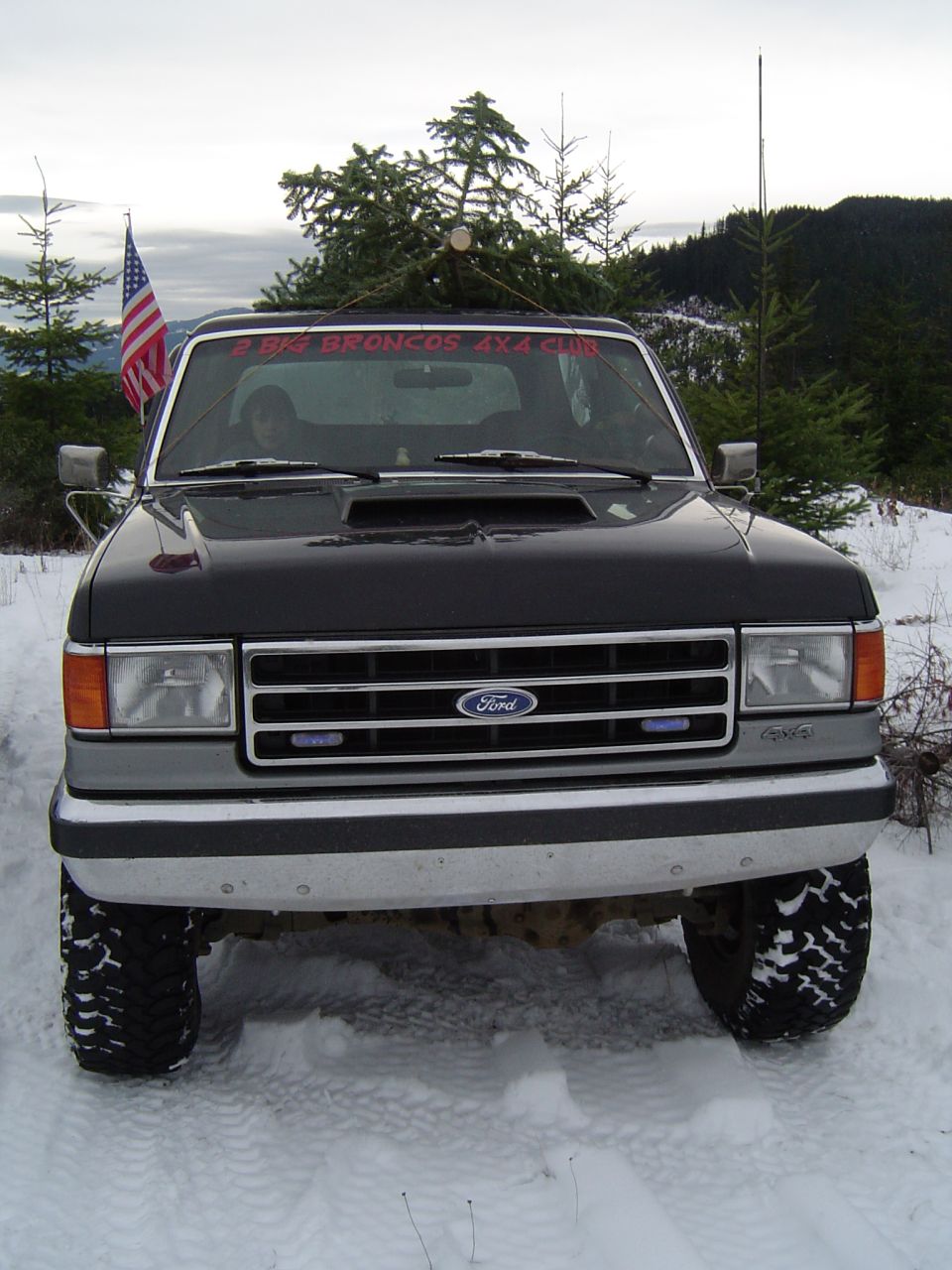 Front of a lifted black-and-silver Ford Bronco on snow with US flag and club banner in the windshield