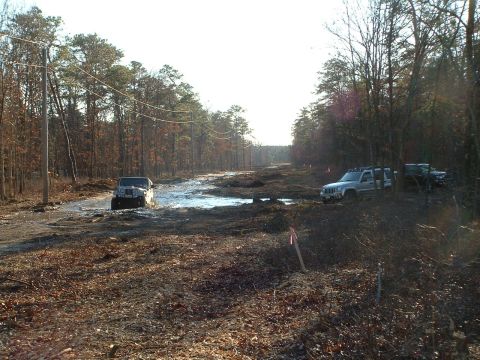 1994 Ford Bronco - Two off-road vehicles navigate a muddy, water-filled path surrounded by trees.