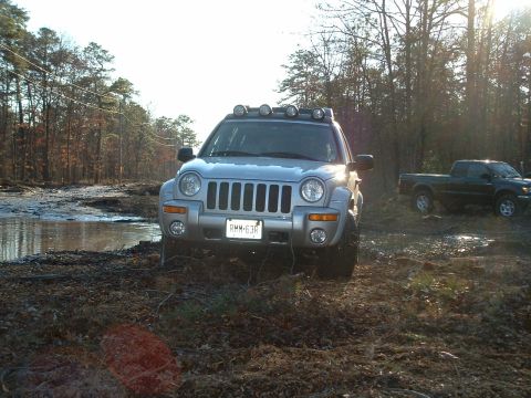 1994 Ford Bronco - A silver Jeep Liberty sits off-road near a muddy area, with a pickup truck visible in the background.