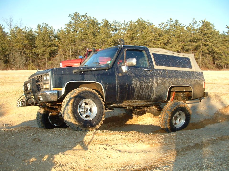 Lifted black SUV with snorkel and off-road tires parked on dirt. Red truck behind it