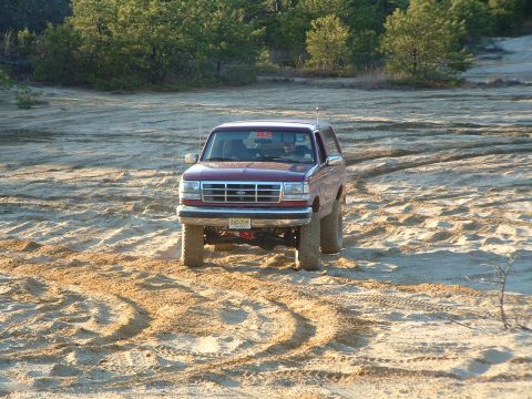 1994 Ford Bronco - A maroon 1994 Ford Bronco navigates a sandy terrain with trees in the background.
