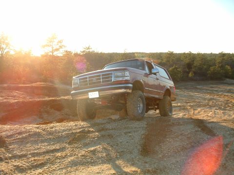 1994 Ford Bronco - A lifted red Ford Bronco navigates a dirt terrain under a bright sun.