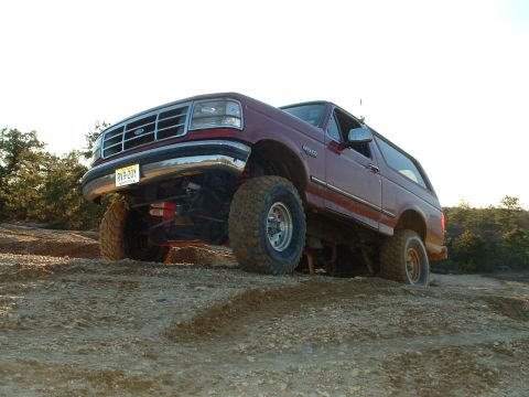 1994 Ford Bronco - This lifted 1994 Ford Bronco navigates a challenging off-road terrain with its front grille and side profile clearly visible.