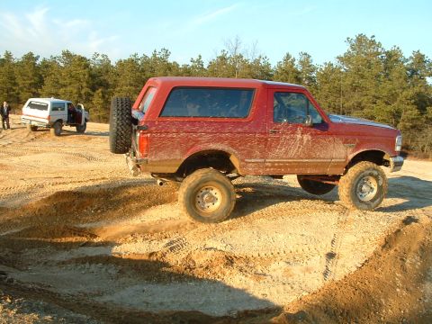1994 Ford Bronco - Two SUVs are seen off-roading on a sandy terrain with trees in the background.