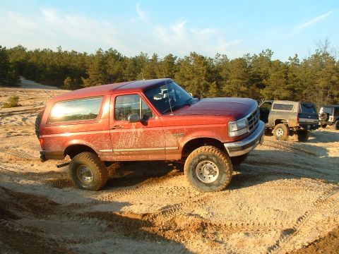 1994 Ford Bronco - Multiple off-road vehicles are parked on a sandy track with a forest in the distance.