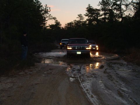 1994 Ford Bronco - A convoy of trucks, including a red 1994 Ford F-150, navigates a muddy trail at dusk.
